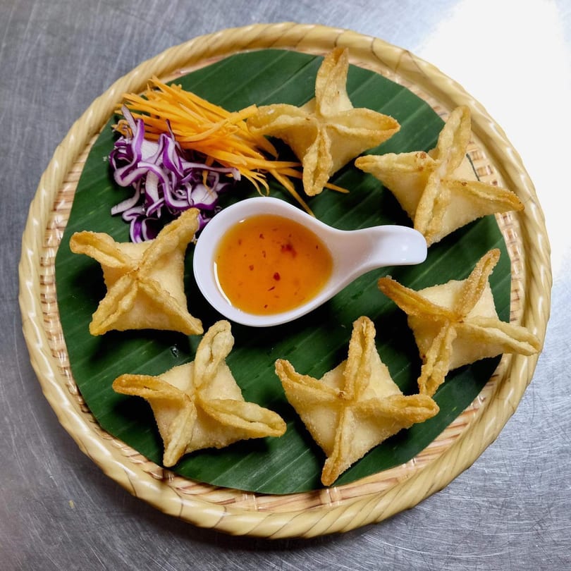 Six golden-fried crab rangoons with sweet chili sauce, shredded carrots, and red cabbage on a banana leaf.