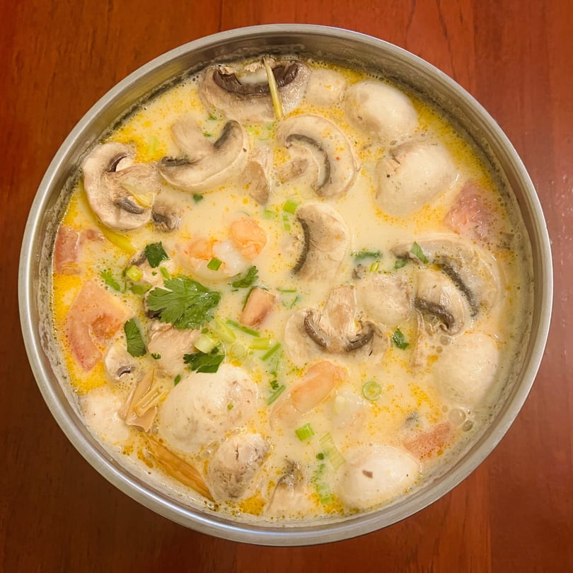 Close-up of Tom Kha Gai soup with mushrooms, shrimp, and herbs in a metal pot.