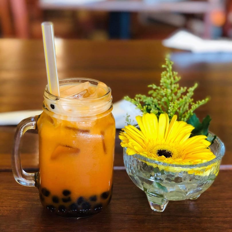 A Thai iced tea with boba in a mason jar next to a yellow gerbera daisy in a glass bowl.