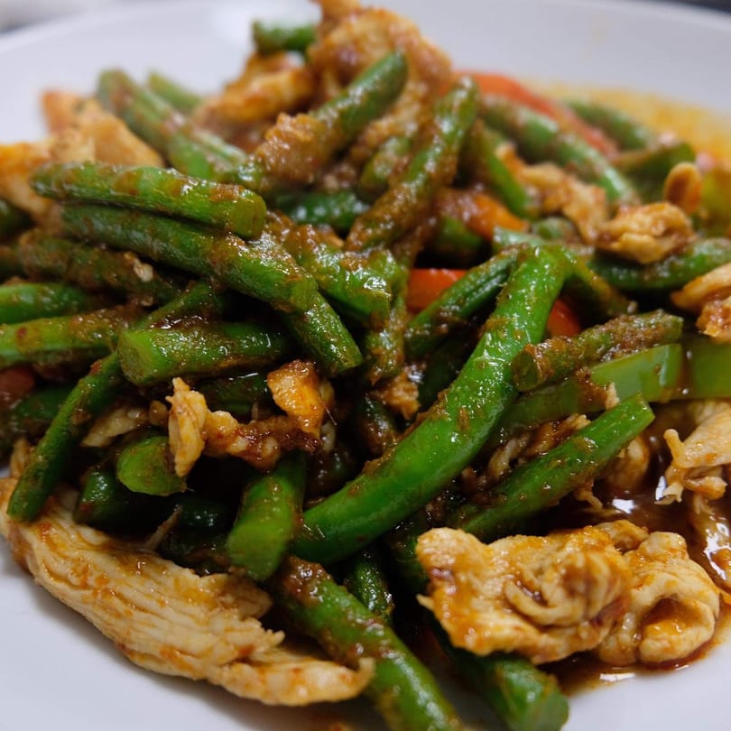 Close-up of Thai chicken and green beans stir-fry with a savory sauce on a white plate.